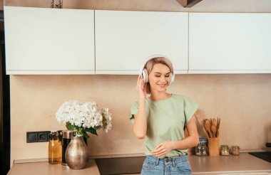 Mutfakta kablosuz kulaklıkla müzik dinleyen mutlu genç bir kadın.