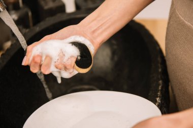 cropped view of woman holding soapy sponge near white plate clipart