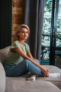 Çekici, düşünceli kadın evdeki pencerenin yanındaki kanepede otururken kameraya bakıyor.