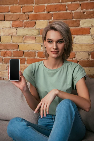 Vacker Ung Kvinna Sitter Soffa Nära Tegelvägg Och Visar Smartphone — Stockfoto