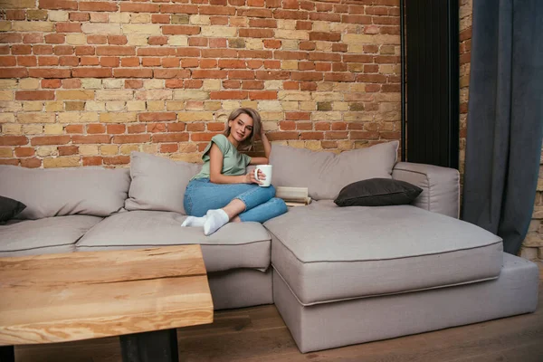 漂亮的年轻女子坐在靠近砖墙的沙发上喝茶 — 图库照片