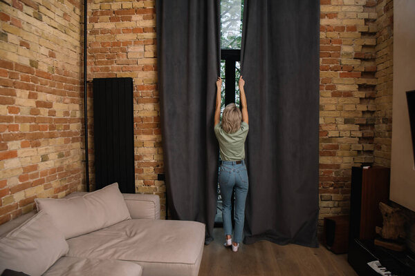 back view of young woman in blue jeans opening window curtains in bedroom