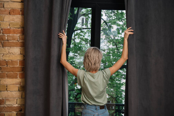back view of young woman opening dark curtains on window at home