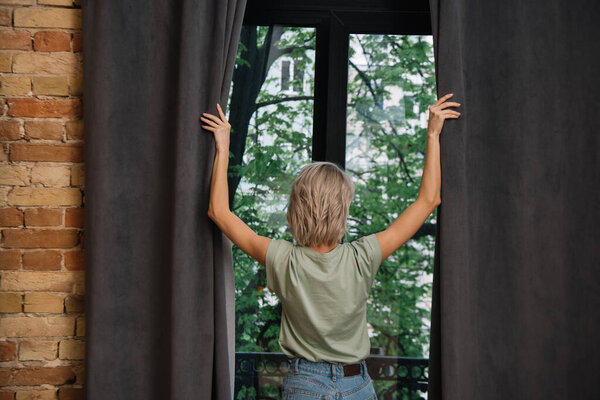 Вид сзади на молодую женщину, открывающую темные шторы на окне дома
