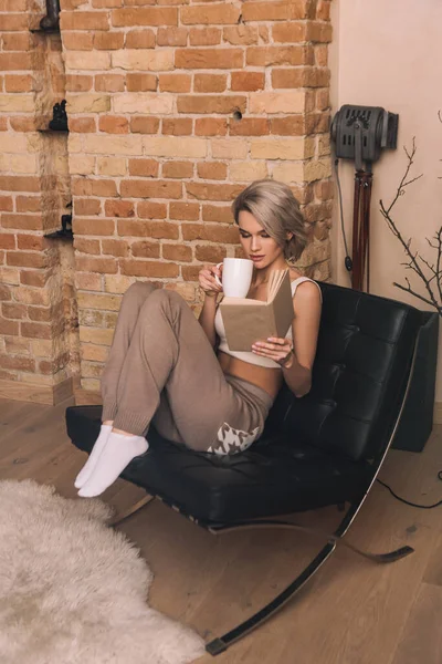 Mulher Jovem Concentrado Leitura Livro Segurando Xícara Chá Enquanto Sentado — Fotografia de Stock