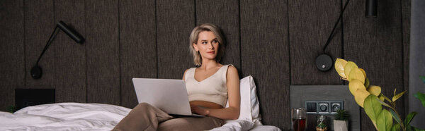 горизонтальное изображение привлекательной молодой женщины, смотрящей в сторону при использовании ноутбука в спальне

