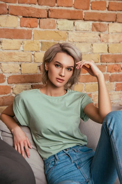 attractive, confident woman sitting on sofa and looking at camera while holding hand near head