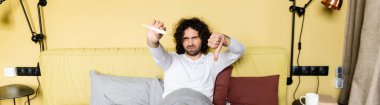 Hoşnutsuz adamın yatay görüntüsü başparmağı aşağıda ve yatakta termometreye bakıyor.