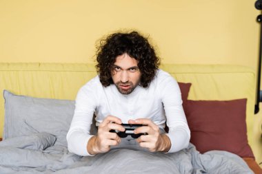 KYIV, UKRAINE - 25 Nisan 2020: konsantre, kıvırcık adam yatak odasında joystick ile video oyunu oynuyor