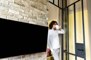 young man in pajamas drinking coffee while standing near blank lcd screen hanging on brick wall clipart