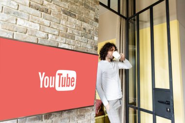 KYIV, UKRAINE - 25 Nisan 2020: Youtube web sitesi ile yeşil lcd ekranının yanında dururken pijamalı genç adam kahve içiyor