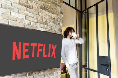KYIV, UKRAINE - APRIL 25, 2020: young man in pajamas drinking coffee while standing near lcd screen with Netflix website clipart