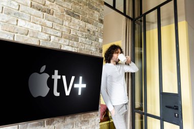 KYIV, UKRAINE - 25 Nisan 2020: Apple TV ile yeşil lcd ekranın yanında dururken pijamalı genç adam kahve içiyor