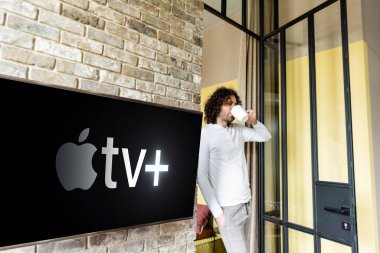KYIV, UKRAINE - APRIL 25, 2020: young man in pajamas drinking coffee while standing near green lcd screen with Apple TV clipart