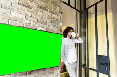 Pijamalı genç adam kahve içerken tuğla duvarda asılı yeşil LCD ekranın yanında duruyor.