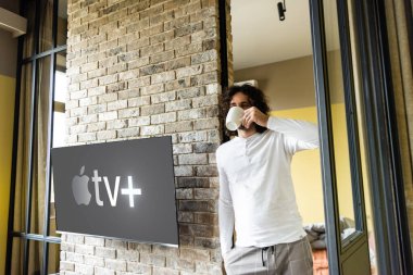KYIV, UKRAINE - 25 Nisan 2020: Apple TV ile Lcd ekranının yanında dururken pijamalı kahve içen genç adam