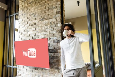 KYIV, UKRAINE - 25 Nisan 2020: Youtube web sitesi ile lcd ekranının yanında dururken pijamalı kahve içen genç adam