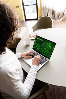Serbest çalışanın, kahve fincanının yanında online ticaret grafikleri ve tostlu tabağı olan dizüstü bilgisayarla yüksek açılı görüntüsü