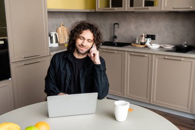 Mutfakta kahve fincanının yanında dizüstü bilgisayar kullanırken akıllı telefondan konuşan serbest çalışan gülümseyen biri.