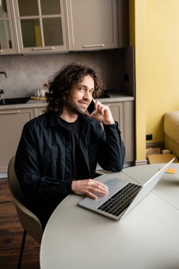 Mutfakta bilgisayarın yanında akıllı telefonla konuşurken kameraya gülümseyen mutlu serbest yazar.