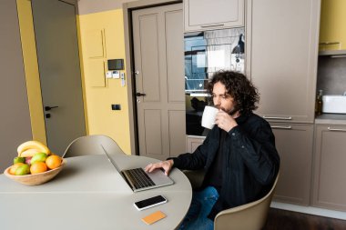 Genç serbest çalışan kahve içerken akıllı telefonun yanında bilgisayarla boş ekran, kredi kartı ve taze meyve kullanıyor.