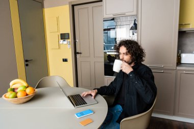 KYIV, UKRAINE - 25 Nisan 2020: Skype uygulaması, kredi kartı ve taze meyve ile akıllı telefon yanında dizüstü bilgisayar kullanarak kahve içen yakışıklı serbest yazar