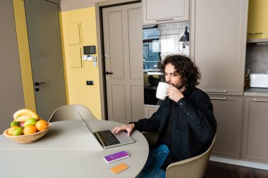 KYIV, UKRAINE - 25 Nisan 2020: Instagram uygulaması, kredi kartı ve taze meyve ile akıllı telefon yanında laptop kullanırken kahve içen yakışıklı serbest yazar
