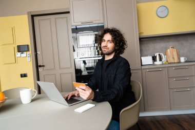 Gülümseyen serbest çalışan, elinde kredi kartı tutarken kameraya bakıyor ve akıllı telefonun yanında beyaz ekranlı bir laptop kullanıyor.