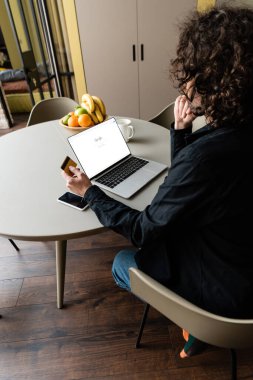 KYIV, UKRAINE - 25 Nisan 2020: Google ekranda, akıllı telefon ve meyvelerle dizüstü bilgisayarda otururken kredi kartı tutan serbest çalışanın arka görünümü