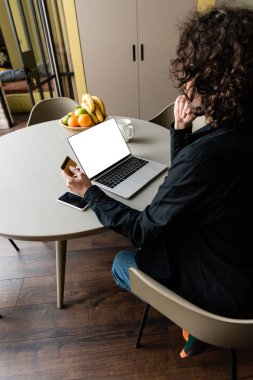 Beyaz ekranlı, akıllı telefonlu ve meyveli dizüstü bilgisayarda otururken kredi kartı tutan serbest çalışanın arka görünümü