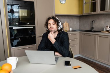Kulaklıklı yakışıklı serbest çalışanın seçici odağı mutfaktaki bilgisayarın yanındaki kredi kartına ve akıllı telefona bakıyor. 