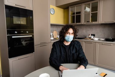 Medikal maskeli bir adam akıllı telefonun yanında dizüstü bilgisayar ve mutfak masasında kredi kartı kullanıyor. 