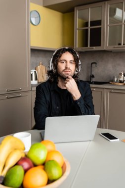 Kulaklıklı bir adamın bilgisayarın yanındaki kameraya, akıllı telefona ve mutfaktaki masada duran meyvelere seçici bir şekilde bakması. 