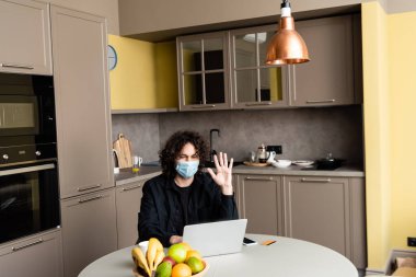 Tıbbi maskeli bir adam bilgisayarında akıllı telefon ve mutfaktaki masada kredi kartı yanında video görüşmesi yapıyor. 