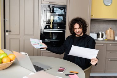 Dizüstü bilgisayarın yanında çizelgeleri olan kafası karışık serbest çalışanın seçici odak noktası ve mutfakta grafikleri olan akıllı telefon. 