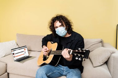 KYIV, UKRAINE - 25 Nisan 2020: Tıp maskeli bir adam oturma odasındaki kanepede SoundCloud web sitesiyle dizüstü bilgisayarın yanında akustik gitar çalıyor