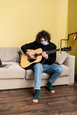 Medikal maskeli bir adam evde Webinar çalarken dizüstü bilgisayar başında akustik gitar çalıyor. 