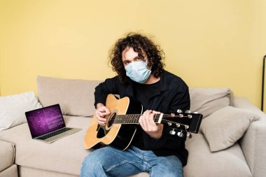 Medikal maskeli kıvırcık adam laptopun yanında akustik gitar çalıyor ve sağlık sitesi kanepede. 