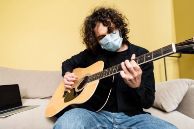 Maskeli bir adam evdeki koltukta dizüstü bilgisayarın yanında akustik gitar çalıyor. 
