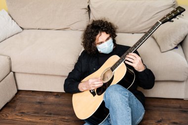 Maskeli bir adam evde yerde akustik gitar çalıyor. 