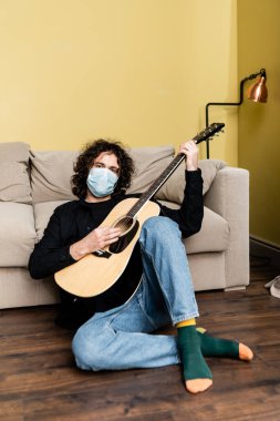 Maskeli bir adam kanepenin yanında akustik gitar çalarken kameraya bakıyor. 