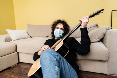 Tıbbi maskeli kıvırcık adam evde yerde otururken akustik gitar çalıyor.