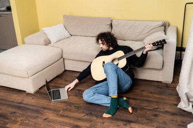Akustik gitar tutan ve evde dizüstü bilgisayar kullanan yakışıklı bir adam. 