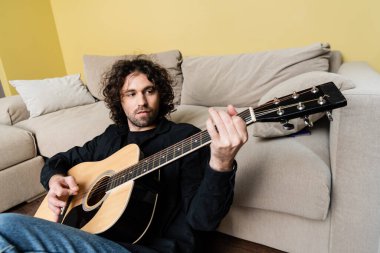 Evde yerde otururken akustik gitar çalan yakışıklı adamın seçici odağı. 