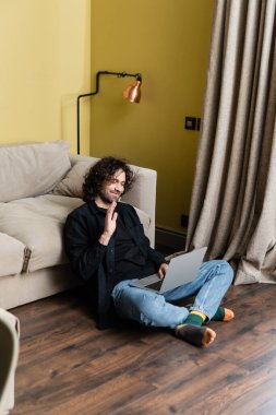 Gülümseyen adamın oturma odasında dizüstü bilgisayarda görüntülü konuşma yapması seçici odak noktası.