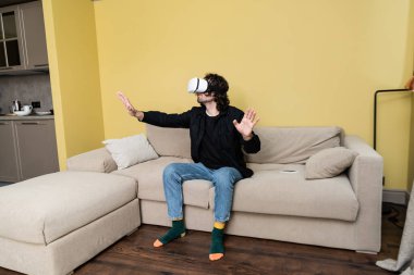 Curly man using virtual reality headset near smartphone on couch at home