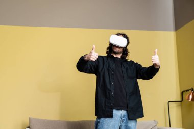 VR kulaklıklı kıvırcık adam oturma odasında baş parmağını gösteriyor.