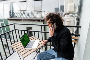 Man talking on smartphone near laptop with online trade website on balcony  clipart