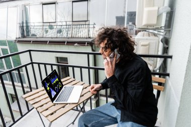 Akıllı telefondan konuşan bir adam dizüstü bilgisayarın yanında, balkonda çizelgeleri olan bir adam. 
