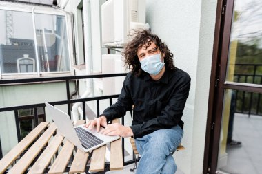 Maskeli bir adam balkonda dizüstü bilgisayar kullanıyor. 